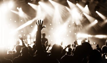 Fotografia em tons de sépia de uma plateia em um show. O público aparece em silhueta contra as luzes brilhantes do palco. Em destaque, no centro, uma mão está erguida para o alto. O fundo está desfocado com vários pontos de luz.